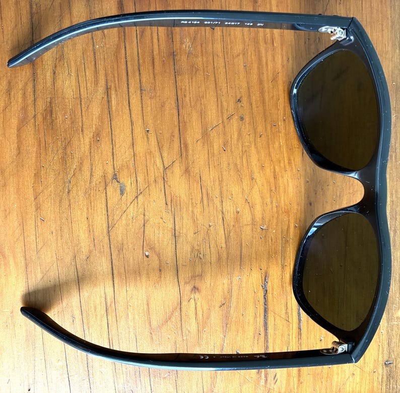 Puede incluir: Gafas de sol negras con lentes oscuras. Las gafas tienen una montura rectangular. Las patillas de las gafas son curvas y se extienden hacia afuera. Sobre un fondo de madera.