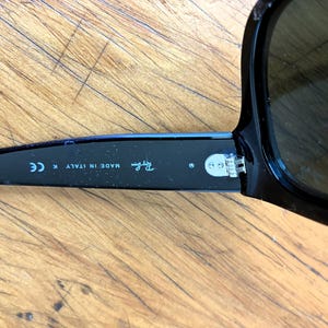 Puede incluir: Gafas de sol negras con lentes oscuras. La patilla izquierda de las gafas muestra el logotipo de Ray-Ban y el texto "MADE IN ITALY K CE". Las gafas est&aacute;n sobre una superficie de madera.