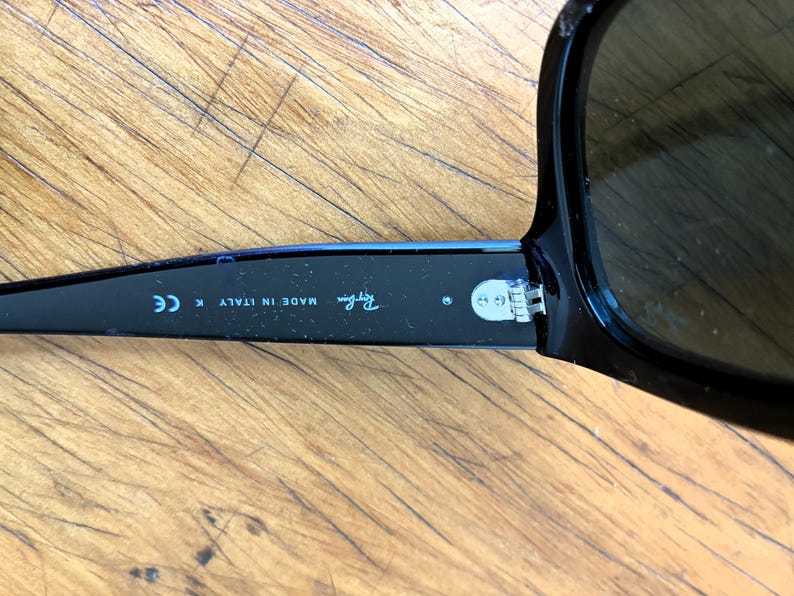 Puede incluir: Gafas de sol negras con lentes oscuras. La patilla izquierda de las gafas muestra el logotipo de Ray-Ban y el texto "MADE IN ITALY K CE". Las gafas est&aacute;n sobre una superficie de madera.