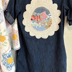 May include: A dark blue denim shirt featuring a large, circular, cream-colored crocheted doily appliqué on the back. The center of the doily contains a patchwork design of various floral and patterned fabric scraps, accented by two small buttons.