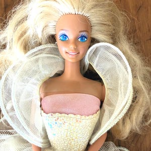 May include: A vintage doll with long, wavy blonde hair and bright blue eyes. The doll wears a pink strapless top and a white and iridescent gown with a sheer, wing-like accent. The doll is a collectible item.