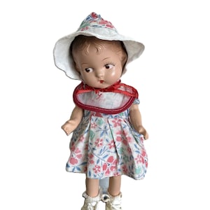 May include: A vintage doll wearing a floral print dress with a matching hat and bib. The doll has painted facial features, including rosy cheeks and red lips. The doll is wearing white shoes. The doll is approximately 10 inches tall.