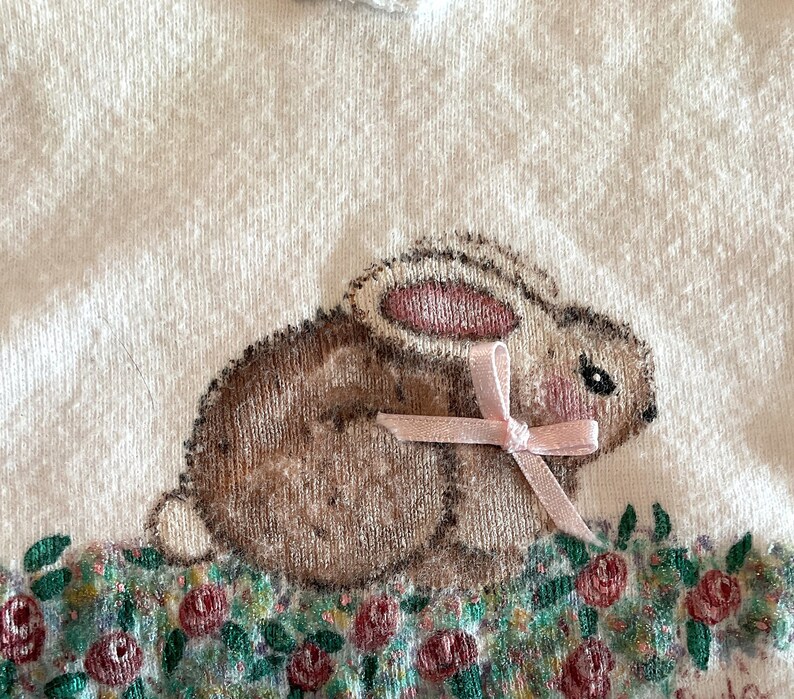 Puede incluir: Tela blanca con una ilustraci&oacute;n detallada de un conejito marr&oacute;n con detalles rosas y un lazo rosa. El conejito est&aacute; sentado en un lecho de flores con detalles rojos y verdes. El dise&ntilde;o est&aacute; centrado en la tela.