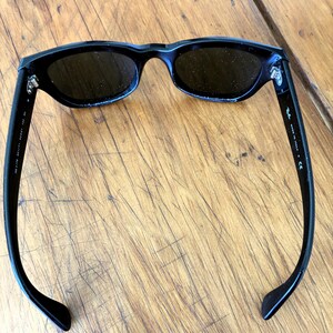 Puede incluir: Gafas de sol negras con lentes oscuras. Las gafas tienen una montura rectangular elegante y patillas curvas. El nombre de la marca es visible en la patilla. Las gafas de sol est&aacute;n sobre una superficie de madera.