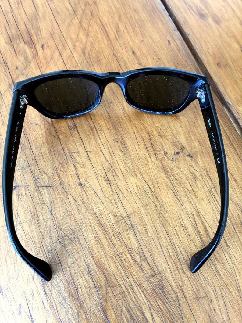 Puede incluir: Gafas de sol negras con lentes oscuras. Las gafas tienen una montura rectangular elegante y patillas curvas. El nombre de la marca es visible en la patilla. Las gafas de sol est&aacute;n sobre una superficie de madera.