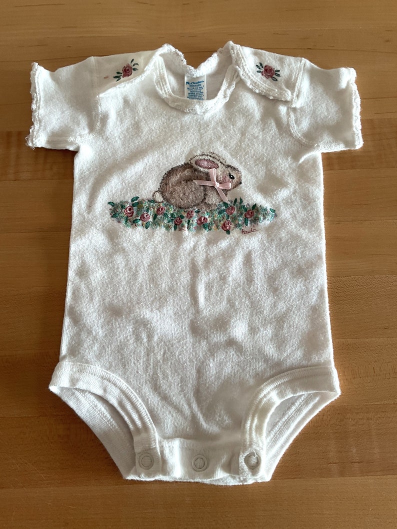Puede incluir: Body de beb&eacute; blanco con bordado de conejito y flores. El body presenta un conejito marr&oacute;n con un lazo rosa, rodeado de flores rosas y verdes. El body de manga corta tiene cierres de presi&oacute;n y bordados florales en los hombros.