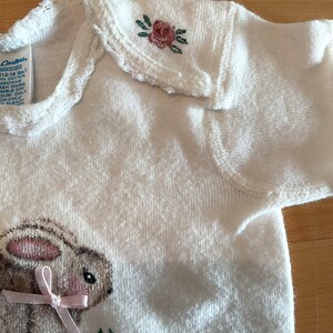 Puede incluir: Body de beb&eacute; blanco con bordado de un conejito y flores. El conejito tiene un lazo rosa y est&aacute; rodeado de flores. El cuello tiene un borde festoneado y una peque&ntilde;a rosa bordada. El body es de algod&oacute;n.