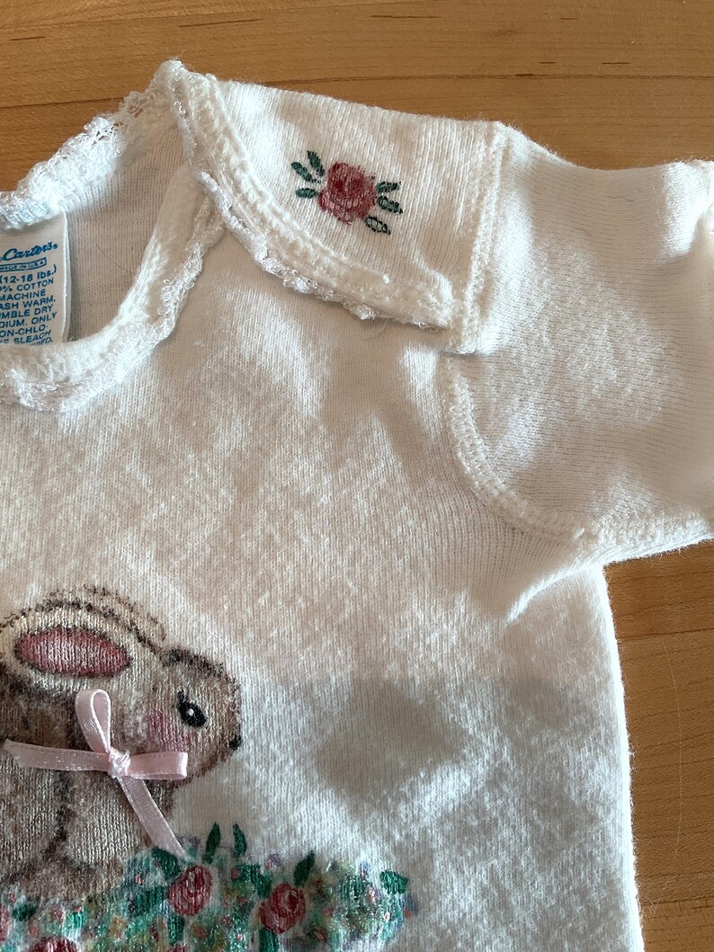 Puede incluir: Body de beb&eacute; blanco con bordado de un conejito y flores. El conejito tiene un lazo rosa y est&aacute; rodeado de flores. El cuello tiene un borde festoneado y una peque&ntilde;a rosa bordada. El body es de algod&oacute;n.