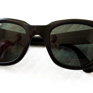 Puede incluir: Gafas de sol negras con lentes oscuras. Las gafas tienen una montura gruesa y cuadrada, y el logotipo de Ray-Ban es visible en la lente superior izquierda. Las lentes reflejan el entorno. Las gafas est&aacute;n sobre una superficie blanca.