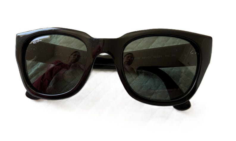 Puede incluir: Gafas de sol negras con lentes oscuras. Las gafas tienen una montura gruesa y cuadrada, y el logotipo de Ray-Ban es visible en la lente superior izquierda. Las lentes reflejan el entorno. Las gafas est&aacute;n sobre una superficie blanca.