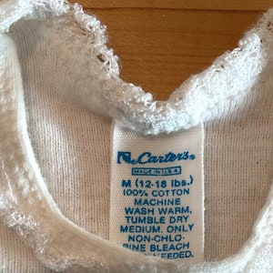 Puede incluir: Primer plano de una camiseta interior blanca para beb&eacute; con una etiqueta de Carter's. La etiqueta indica la talla M (5,4-8,2 kg) y est&aacute; hecha de 100% algod&oacute;n. La camiseta tiene un ribete blanco y con volantes alrededor del cuello.