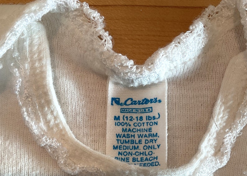 Puede incluir: Primer plano de una camiseta interior blanca para beb&eacute; con una etiqueta de Carter's. La etiqueta indica la talla M (5,4-8,2 kg) y est&aacute; hecha de 100% algod&oacute;n. La camiseta tiene un ribete blanco y con volantes alrededor del cuello.