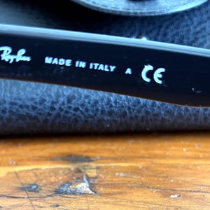 Puede incluir: Primer plano de una patilla de gafas negra con el logotipo de Ray-Ban y el texto "MADE IN ITALY A CE" en blanco. La patilla descansa sobre un estuche de cuero azul oscuro, con una superficie de madera en primer plano.