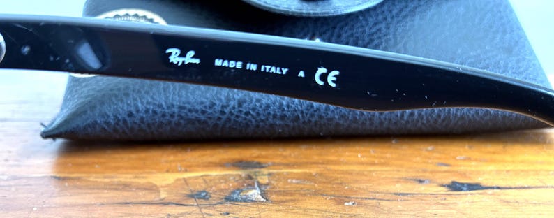 Puede incluir: Primer plano de una patilla de gafas negra con el logotipo de Ray-Ban y el texto "MADE IN ITALY A CE" en blanco. La patilla descansa sobre un estuche de cuero azul oscuro, con una superficie de madera en primer plano.