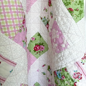 May include: A quilted blanket featuring a patchwork design of diamond-shaped fabric pieces. The pattern includes light green squares with pink and red rose floral prints, pink and white plaid, pink polka dots, and white fabric with small floral motifs.