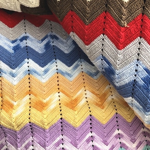 May include: A colorful, handmade crochet blanket with a chevron pattern. The blanket features a variety of colors, including cream, brown, red, blue, yellow, purple, and teal. The blanket is folded, showcasing the different color sections.