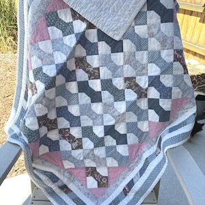 May include: A patchwork quilt featuring a repeating geometric pattern in shades of dusty pink, slate blue, charcoal, and white. The fabric squares display various floral and small-scale prints, finished with a white border and quilted texture.