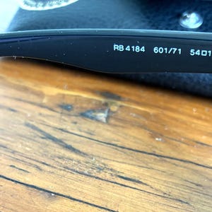 Puede incluir: Primer plano de la patilla de unas gafas de sol con montura negra. El texto "RB4184 601/71 54017 145 3N" est&aacute; impreso en blanco en el interior de la patilla. Las gafas de sol est&aacute;n sobre una superficie de madera.
