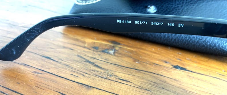 Puede incluir: Primer plano de la patilla de unas gafas de sol con montura negra. El texto "RB4184 601/71 54017 145 3N" est&aacute; impreso en blanco en el interior de la patilla. Las gafas de sol est&aacute;n sobre una superficie de madera.