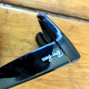 Puede incluir: Primer plano de gafas de sol negras con el logotipo de Ray-Ban en escritura blanca. Las gafas de sol tienen un acabado brillante y un dise&ntilde;o cl&aacute;sico. La imagen est&aacute; tomada en &aacute;ngulo, mostrando la montura y parte de la lente.