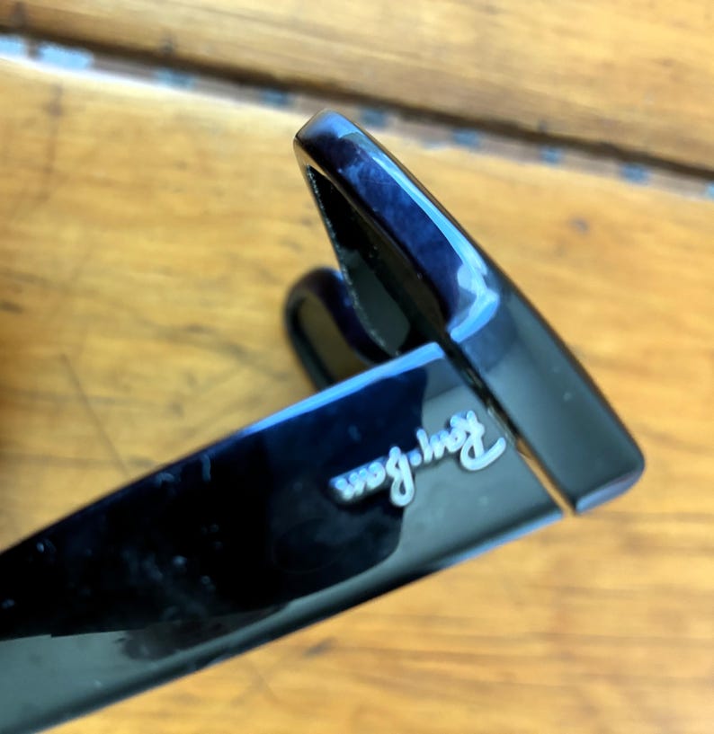 Puede incluir: Primer plano de gafas de sol negras con el logotipo de Ray-Ban en escritura blanca. Las gafas de sol tienen un acabado brillante y un dise&ntilde;o cl&aacute;sico. La imagen est&aacute; tomada en &aacute;ngulo, mostrando la montura y parte de la lente.