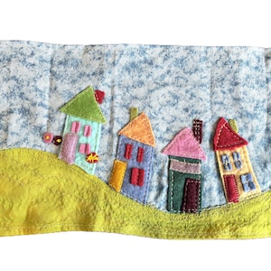 May include: A rectangular fabric wall hanging featuring a quilted blue and white mottled sky background above a textured yellow hill. Four colorful felt houses are appliqued across the scene, each with distinct roof colors and stitched details.