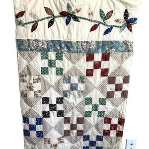 May include: A patchwork quilt featuring a cream-colored top border with an appliqued vine of multi-colored fabric leaves. Below this, a floral strip separates the top from a grid of square blocks in various patterns, including gingham and floral prints.