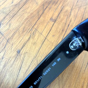 Puede incluir: Primer plano de una patilla de gafas de sol negra con el texto "RB 4178 601/71 52021 140 3N". La patilla es de material negro brillante. El fondo es una superficie de madera marr&oacute;n claro.