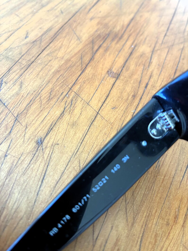 Puede incluir: Primer plano de una patilla de gafas de sol negra con el texto "RB 4178 601/71 52021 140 3N". La patilla es de material negro brillante. El fondo es una superficie de madera marr&oacute;n claro.