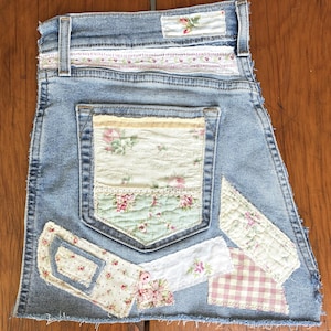 May include: A light blue denim skirt featuring a frayed hem and various fabric patches. The back pocket and waistband are decorated with floral and gingham patterned fabric scraps, lace trim, and a small pink button on the lower left side.