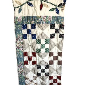 May include: A rectangular patchwork quilt featuring a cream-colored top border with appliquéd fabric leaves in various colors. The main body consists of alternating square blocks in floral, checkered, and solid patterns in blue, red, and white.