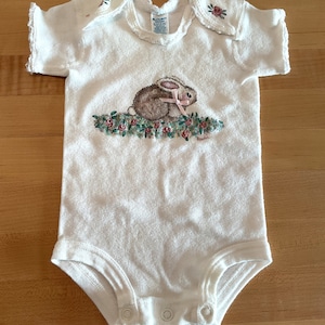 Puede incluir: Body de beb&eacute; blanco con una ilustraci&oacute;n de un conejito. El conejo es marr&oacute;n con orejas rosas y un lazo rosa, sentado sobre un lecho de flores rosas y verdes. El body tiene mangas cortas y corchetes en la parte inferior.
