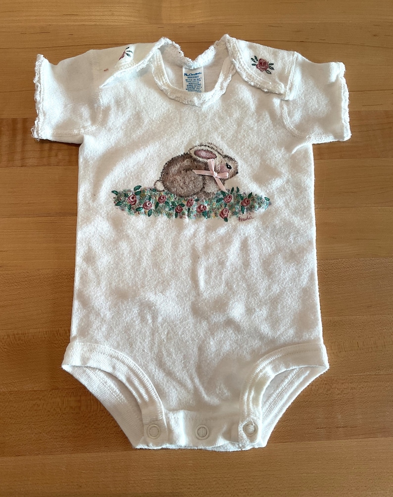 Puede incluir: Body de beb&eacute; blanco con una ilustraci&oacute;n de un conejito. El conejo es marr&oacute;n con orejas rosas y un lazo rosa, sentado sobre un lecho de flores rosas y verdes. El body tiene mangas cortas y corchetes en la parte inferior.