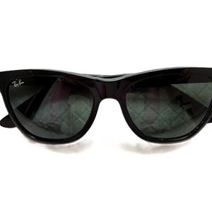 Puede incluir: Gafas de sol negras con montura de ojo de gato. Las lentes son oscuras y el logotipo de Ray-Ban es visible en la lente izquierda. Fondo blanco.