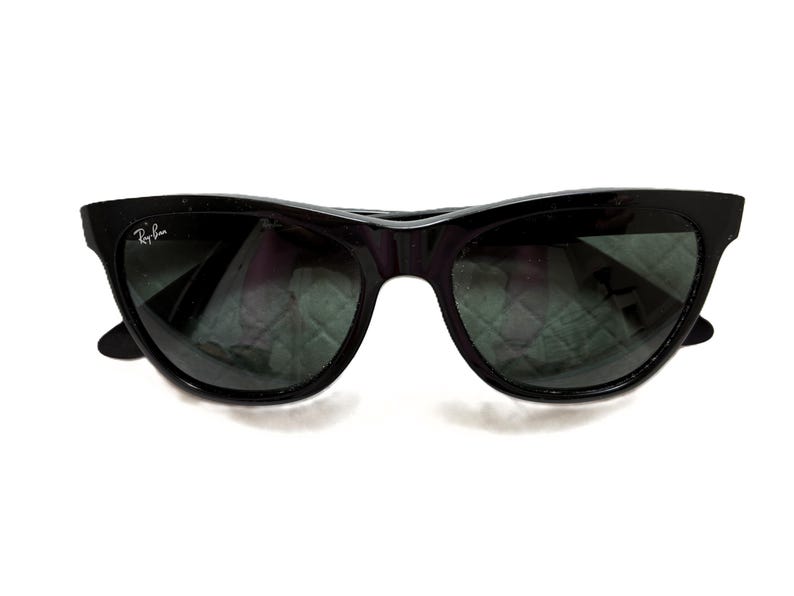 Puede incluir: Gafas de sol negras con montura de ojo de gato. Las lentes son oscuras y el logotipo de Ray-Ban es visible en la lente izquierda. Fondo blanco.