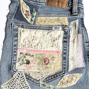 May include: Light blue denim jeans featuring a brown leather Signature Levi Strauss patch. The back pocket and surrounding areas are decorated with various fabric patches, including floral patterns, cream-colored lace, and quilted textile segments.
