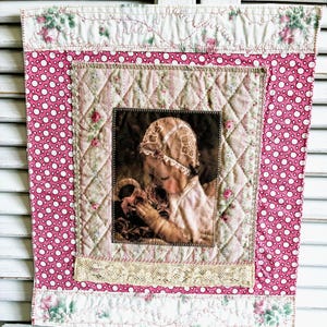 May include: A rectangular fabric wall hanging featuring a central photographic print of a child wearing a lace bonnet and holding roses. The print is framed by quilted pink and white striped fabric, a pink polka dot border, and floral trim.
