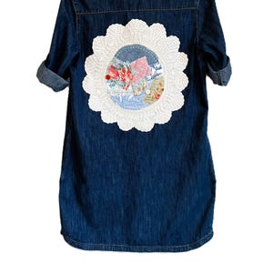 May include: A dark blue denim shirt with rolled sleeves featuring a large, circular white crocheted doily appliqué on the back. The center of the doily contains a collage of floral and patterned fabric patches in shades of red, pink, and blue.