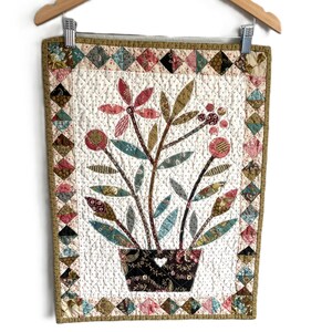 May include: A rectangular quilted wall hanging featuring a central applique of a dark brown pot with a heart cutout, containing stylized flowers and leaves in shades of pink, teal, and olive. The border consists of small, multi-colored fabric squares.