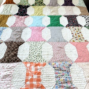 May include: A colorful quilt with a repeating spool design. The quilt features various patterned fabrics in shades of pink, green, blue, orange, and brown, set against a white background. The quilt is quilted with a diamond pattern.