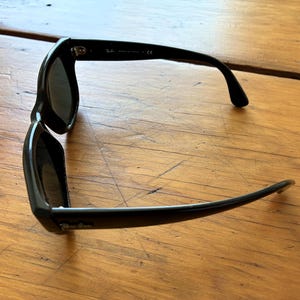 Puede incluir: Gafas de sol negras con lentes oscuras. Las gafas tienen una montura rectangular y patillas curvas. El nombre de la marca es visible en la patilla. Las gafas descansan sobre una superficie de madera.
