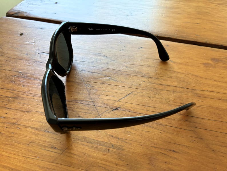 Puede incluir: Gafas de sol negras con lentes oscuras. Las gafas tienen una montura rectangular y patillas curvas. El nombre de la marca es visible en la patilla. Las gafas descansan sobre una superficie de madera.
