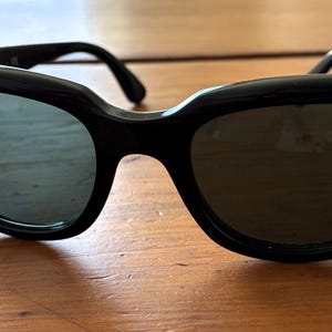 Puede incluir: Gafas de sol negras de ojo de gato con lentes oscuras. Las gafas tienen una montura negra gruesa y brillante y el logotipo de Ray-Ban es visible en la lente izquierda. Las gafas descansan sobre una superficie de madera.