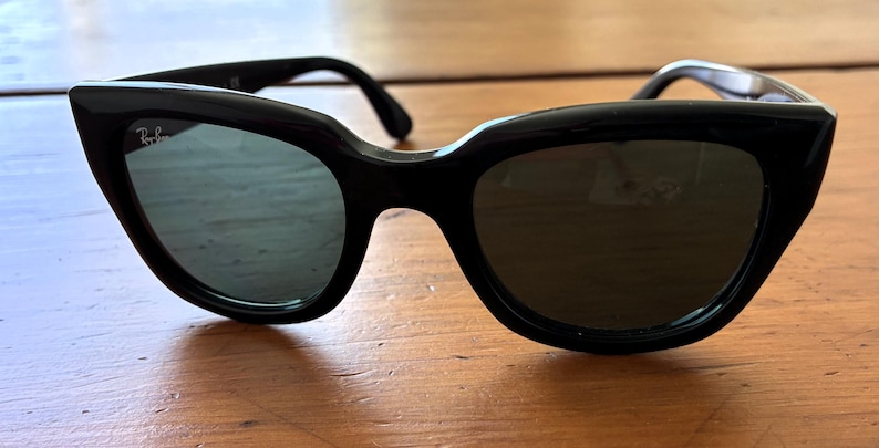 Puede incluir: Gafas de sol negras de ojo de gato con lentes oscuras. Las gafas tienen una montura negra gruesa y brillante y el logotipo de Ray-Ban es visible en la lente izquierda. Las gafas descansan sobre una superficie de madera.