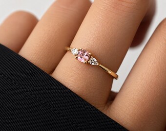 Puede incluir: Un anillo de oro con una piedra preciosa rosa central de corte cojín, flanqueada por dos piedras blancas transparentes más pequeñas de corte en forma de pera. La banda es fina y pulida, con un delicado diseño de tres piedras.