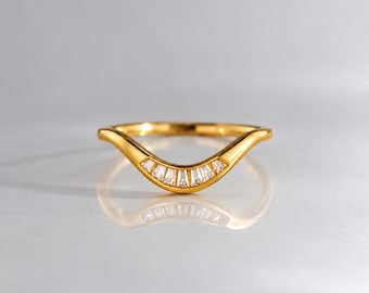 Pode incluir: Um anel dourado curvo com um design de banda em forma de V. O centro da banda é cravejado com uma fileira de sete pequenas pedras transparentes de corte baguette, dispostas em leque para criar um arco subtil na parte frontal do anel.