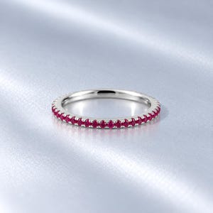 May include: A thin silver-toned band ring featuring a continuous row of small, round, vibrant red gemstones set along the outer circumference. The metal has a polished, reflective finish, and the stones are evenly spaced in a shared prong setting.