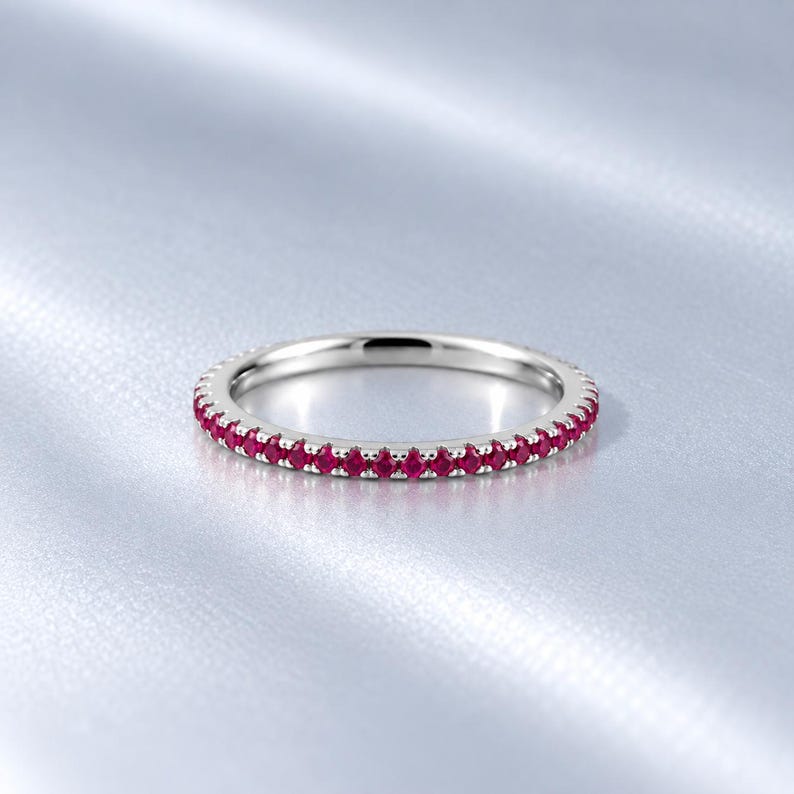 May include: A thin silver-toned band ring featuring a continuous row of small, round, vibrant red gemstones set along the outer circumference. The metal has a polished, reflective finish, and the stones are evenly spaced in a shared prong setting.