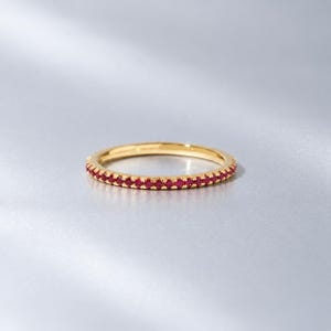 May include: A thin gold band ring featuring a continuous row of small, round, vibrant red gemstones set along the entire outer circumference. The metal has a polished, reflective finish, and the stones are uniform in size and colour throughout.