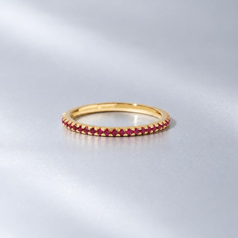 May include: A thin gold band ring featuring a continuous row of small, round, vibrant red gemstones set along the entire outer circumference. The metal has a polished, reflective finish, and the stones are uniform in size and colour throughout.